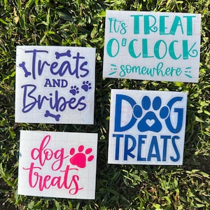 May include: Four square white decals with text related to dog treats. The decals are on a green grassy surface. The text includes "Treats and Bribes", "It's Treat O'Clock somewhere", "dog treats", and "Dog Treats".