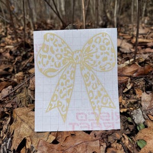 Peut inclure: Un autocollant adh&eacute;sif jaune clair en forme de n&oelig;ud avec un motif l&eacute;opard. L'autocollant est appliqu&eacute; sur une feuille de support rectangulaire blanche pr&eacute;sentant un l&eacute;ger motif quadrill&eacute; et du texte rouge partiellement visible en bas.