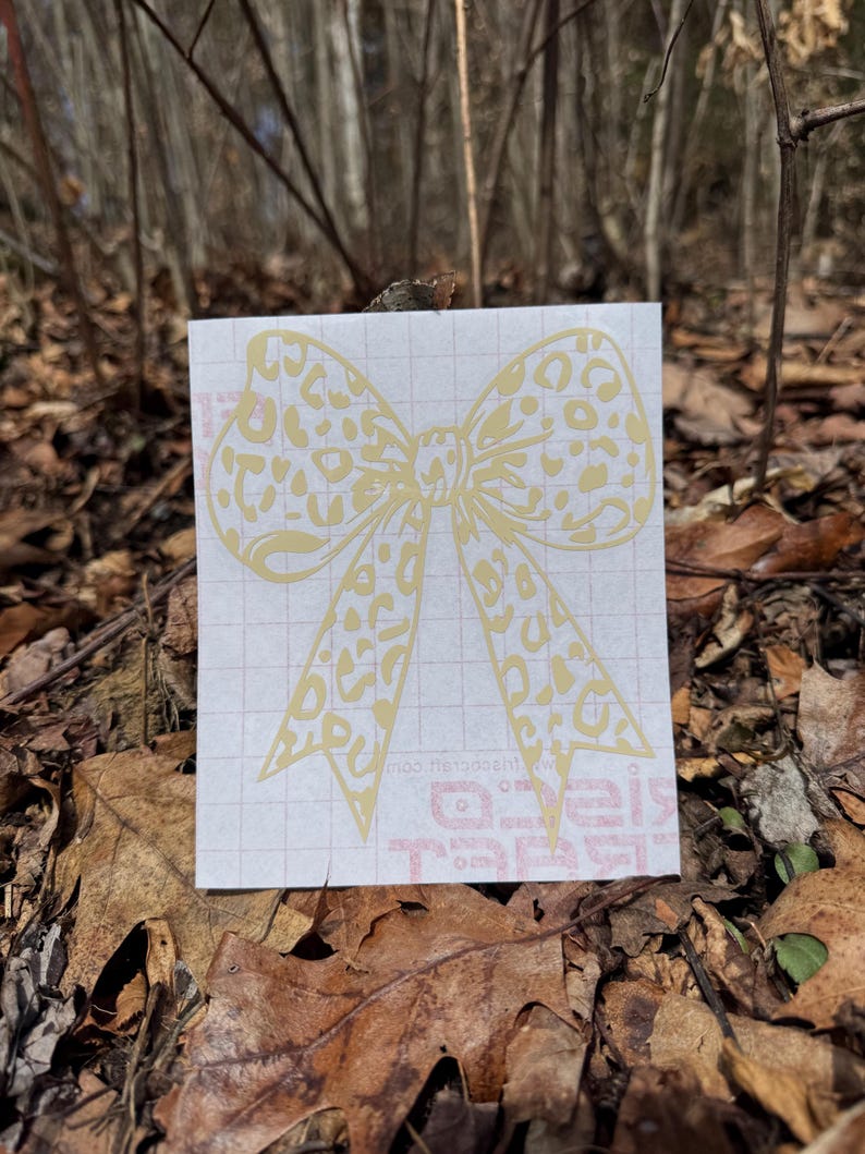 Peut inclure: Un autocollant adh&eacute;sif jaune clair en forme de n&oelig;ud avec un motif l&eacute;opard. L'autocollant est appliqu&eacute; sur une feuille de support rectangulaire blanche pr&eacute;sentant un l&eacute;ger motif quadrill&eacute; et du texte rouge partiellement visible en bas.