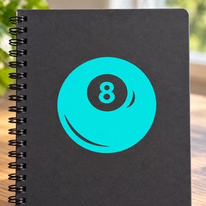 May include: A black spiral-bound notebook featuring a large, bright cyan graphic of an eight ball on the front cover. The design displays a solid cyan circle with a dark gray center containing the white number eight and a curved highlight detail.