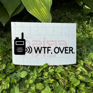 Könnte beinhalten: Ein weißer Vinyl-Aufkleber mit einer schwarzen Grafik eines Walkie-Talkies und den Worten "WTF, OVER." Der rechteckige Aufkleber liegt auf einem Bett aus grünem Gras und Blättern.