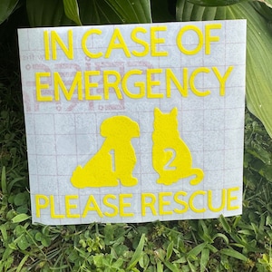 May include: A rectangular sign with yellow text and graphics, reading "IN CASE OF EMERGENCY PLEASE RESCUE." Silhouettes of a dog and cat are below the text, with the numbers 1 and 2 next to them. The sign is on a grassy surface.