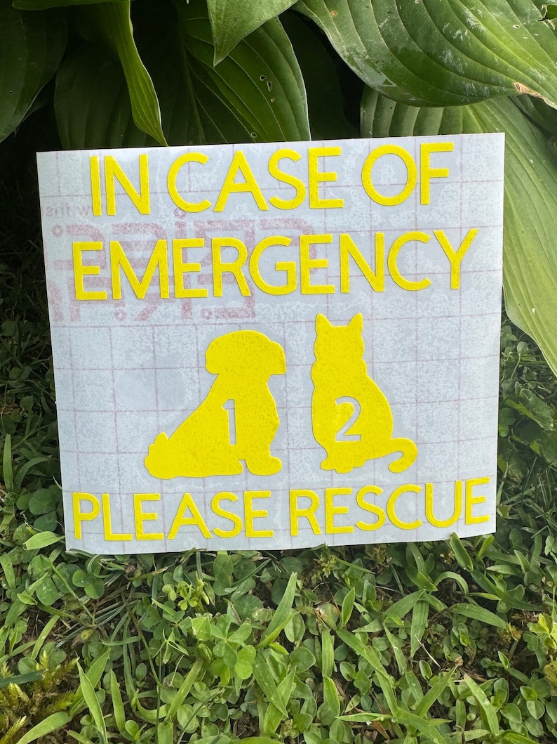 May include: A rectangular sign with yellow text and graphics, reading "IN CASE OF EMERGENCY PLEASE RESCUE." Silhouettes of a dog and cat are below the text, with the numbers 1 and 2 next to them. The sign is on a grassy surface.