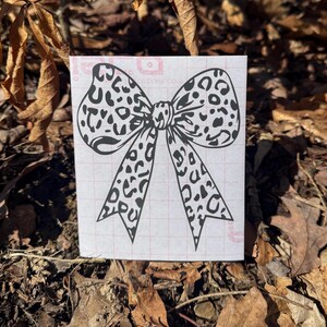 Peut inclure: Une feuille rectangulaire blanche pr&eacute;sentant un motif de n&oelig;ud &agrave; imprim&eacute; l&eacute;opard noir. Le n&oelig;ud poss&egrave;de deux grandes boucles et deux longs pans pointus. La feuille comporte un quadrillage rouge p&acirc;le imprim&eacute; sur la surface, visible derri&egrave;re le motif.