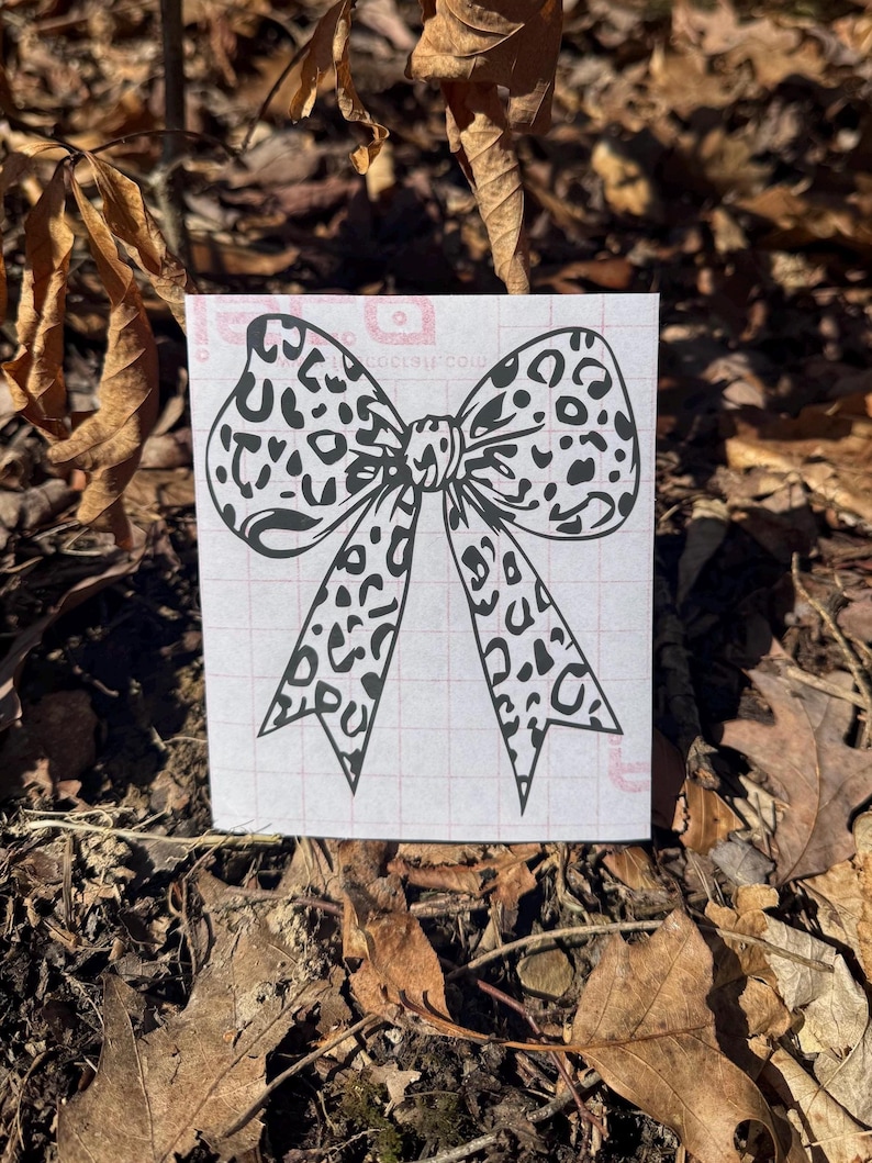 Peut inclure: Une feuille rectangulaire blanche pr&eacute;sentant un motif de n&oelig;ud &agrave; imprim&eacute; l&eacute;opard noir. Le n&oelig;ud poss&egrave;de deux grandes boucles et deux longs pans pointus. La feuille comporte un quadrillage rouge p&acirc;le imprim&eacute; sur la surface, visible derri&egrave;re le motif.