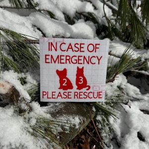 May include: A red and white sticker with the text "IN CASE OF EMERGENCY" and "PLEASE RESCUE". The sticker features silhouettes of two pets, numbered 2 and 3, against a snowy backdrop of pine branches.