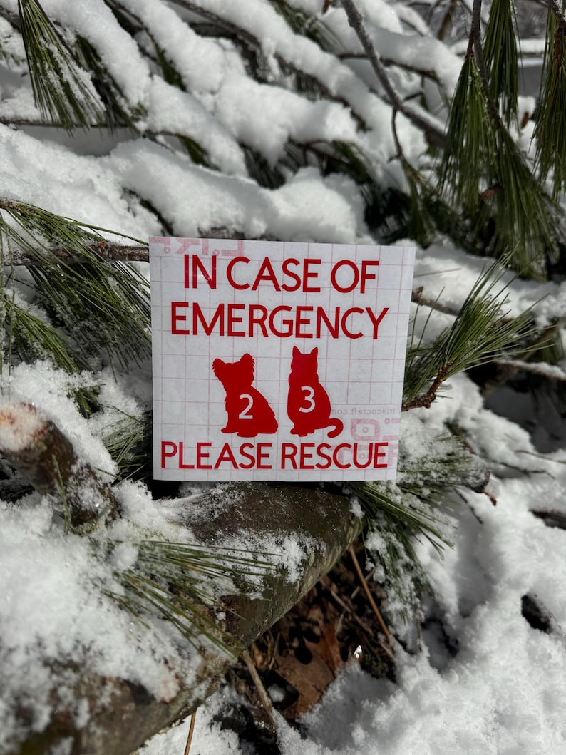 May include: A red and white sticker with the text "IN CASE OF EMERGENCY" and "PLEASE RESCUE". The sticker features silhouettes of two pets, numbered 2 and 3, against a snowy backdrop of pine branches.