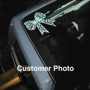 Peut inclure: Un autocollant blanc en forme de n&oelig;ud avec un motif l&eacute;opard appliqu&eacute; sur le coin sup&eacute;rieur d'une vitre de v&eacute;hicule. Le n&oelig;ud pr&eacute;sente un centre nou&eacute; et deux extr&eacute;mit&eacute;s de ruban tombantes, le tout dans un design graphique blanc uni.