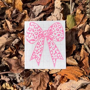 Peut inclure: Un autocollant en vinyle rose vif repr&eacute;sentant un n&oelig;ud avec un motif l&eacute;opard. L'autocollant est appliqu&eacute; sur une feuille de support rectangulaire blanche qui pr&eacute;sente un l&eacute;ger motif de grille grise sur toute sa surface pour l'alignement.