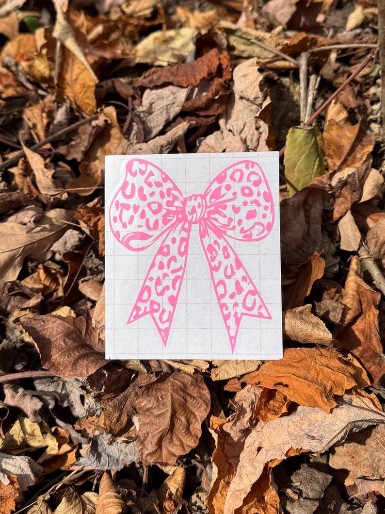Peut inclure: Un autocollant en vinyle rose vif repr&eacute;sentant un n&oelig;ud avec un motif l&eacute;opard. L'autocollant est appliqu&eacute; sur une feuille de support rectangulaire blanche qui pr&eacute;sente un l&eacute;ger motif de grille grise sur toute sa surface pour l'alignement.