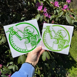 May include: Two square white decals featuring a bright green graphic of a skeleton drinking from a gas pump nozzle. The circular design includes the text GAS GUZZLER in green capital letters. The decals are printed on a grid-lined backing paper.