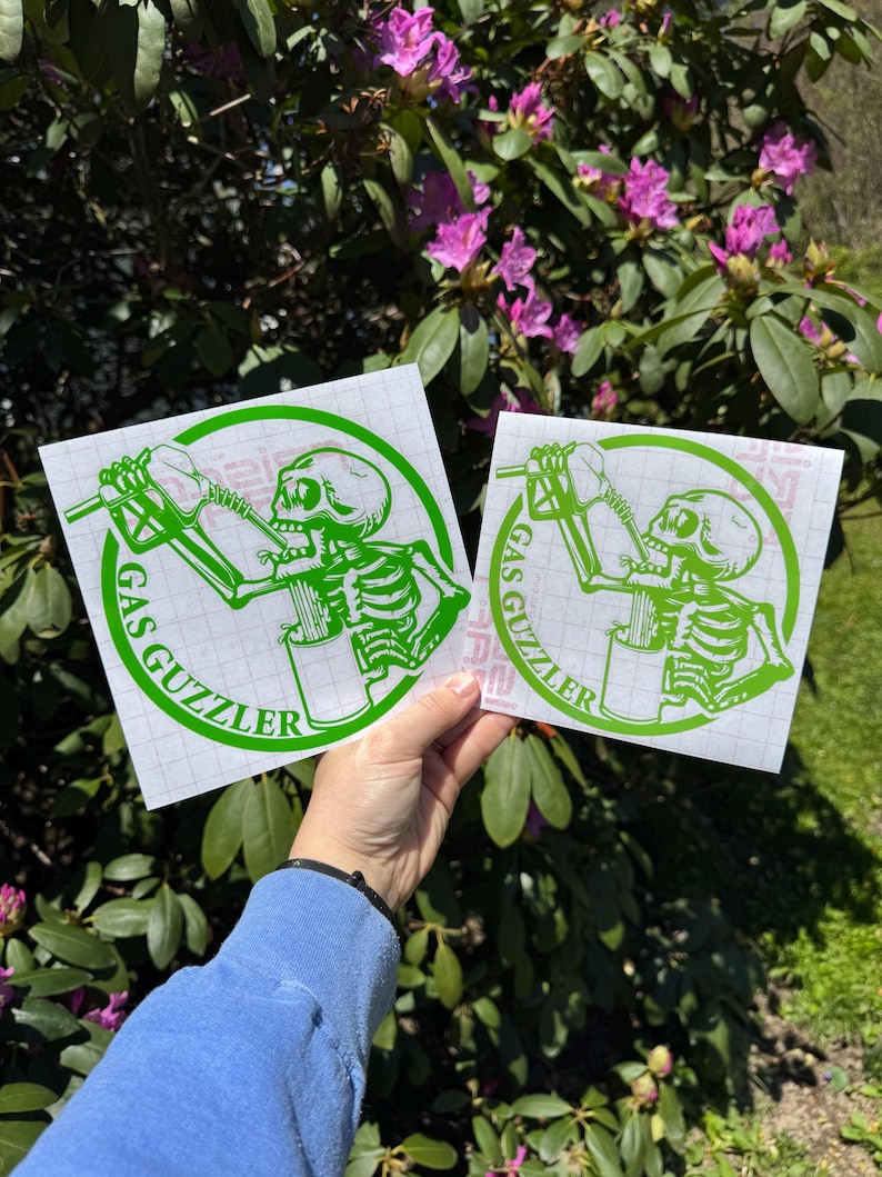 May include: Two square white decals featuring a bright green graphic of a skeleton drinking from a gas pump nozzle. The circular design includes the text GAS GUZZLER in green capital letters. The decals are printed on a grid-lined backing paper.