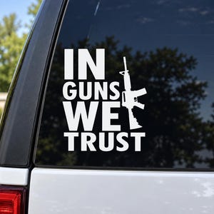 May include: A white vinyl decal applied to a vehicle window featuring the text IN GUNS WE TRUST in a bold, sans-serif font. The letter U in the word GUNS is replaced by a white silhouette of a rifle, positioned vertically to the right of the text.