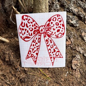 Peut inclure: Un autocollant en vinyle rouge repr&eacute;sentant un n&oelig;ud avec un motif l&eacute;opard. L'autocollant est appliqu&eacute; sur une feuille de support rectangulaire blanche avec un quadrillage l&eacute;ger et du texte partiellement visible en bas, pr&eacute;sent&eacute; comme un graphique adh&eacute;sif plat.