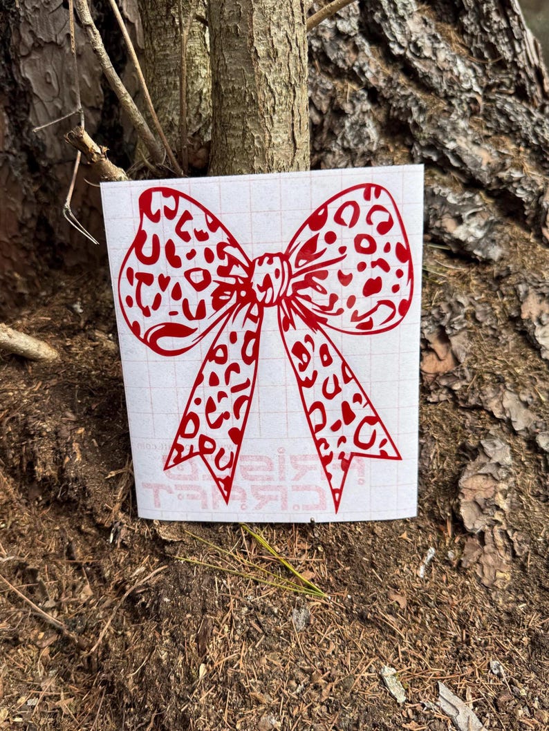 Peut inclure: Un autocollant en vinyle rouge repr&eacute;sentant un n&oelig;ud avec un motif l&eacute;opard. L'autocollant est appliqu&eacute; sur une feuille de support rectangulaire blanche avec un quadrillage l&eacute;ger et du texte partiellement visible en bas, pr&eacute;sent&eacute; comme un graphique adh&eacute;sif plat.