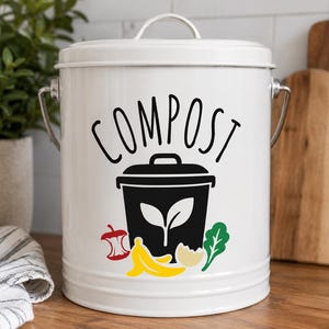 Könnte beinhalten: Ein weißer, zylindrischer Komposteimer aus Metall mit Deckel und silbernem Griff. Auf der Vorderseite befindet sich der schwarze Schriftzug COMPOST über einem schwarzen Mülleimer-Symbol mit weißem Blatt, umgeben von bunten Illustrationen eines roten Apfelgehäuses, gelber Bananenschalen, einer beigen Eierschale und einem grünen Blattgemüse.