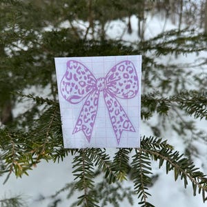 Peut inclure: Un autocollant en vinyle violet clair repr&eacute;sentant un n&oelig;ud avec un motif l&eacute;opard. L'autocollant est appliqu&eacute; sur une feuille de support rectangulaire blanche avec un quadrillage rouge p&acirc;le, con&ccedil;u pour un alignement et une pose faciles.
