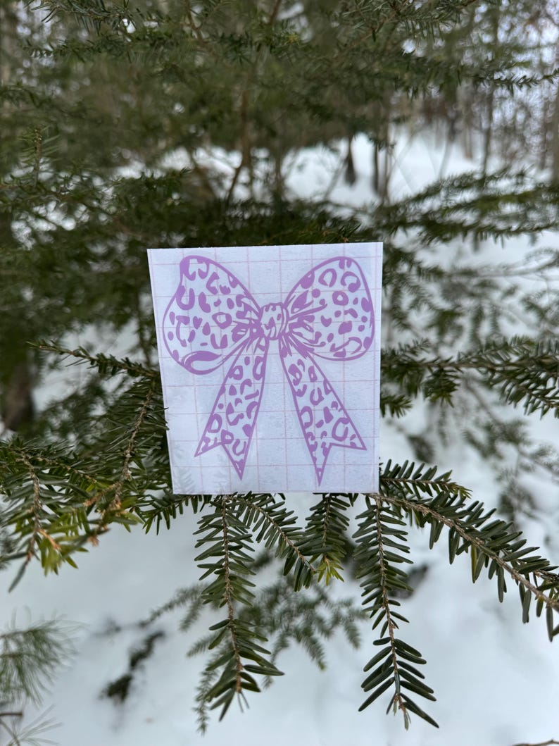Peut inclure: Un autocollant en vinyle violet clair repr&eacute;sentant un n&oelig;ud avec un motif l&eacute;opard. L'autocollant est appliqu&eacute; sur une feuille de support rectangulaire blanche avec un quadrillage rouge p&acirc;le, con&ccedil;u pour un alignement et une pose faciles.