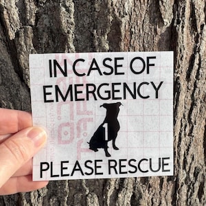 May include: A white square sticker with black text that reads "IN CASE OF EMERGENCY PLEASE RESCUE". A black silhouette of a dog is in the center with the number "1" inside. The sticker is held in front of a tree trunk.