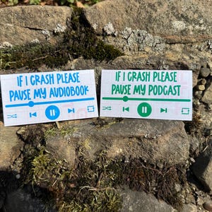 Pode incluir: Dois autocolantes retangulares com o texto "IF I CRASH PLEASE PAUSE MY AUDIOBOOK" e "IF I CRASH PLEASE PAUSE MY PODCAST". Cada autocolante tem um design de leitor multimédia com botões de reprodução, pausa e avanço. O texto e o design são em azul e verde.