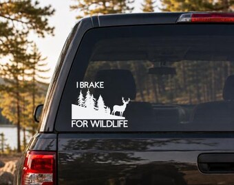 May include: A white vinyl decal applied to a vehicle rear window featuring a silhouette of a forest with pine trees and a standing deer. The text I BRAKE FOR WILDLIFE is printed in a bold sans-serif font above and below the graphic design.