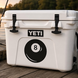 May include: A white YETI cooler featuring a black and white 8-ball graphic on the front. The rectangular cooler has two black rubber T-latches on the lid and the YETI brand name printed in white letters on a black rectangular background.