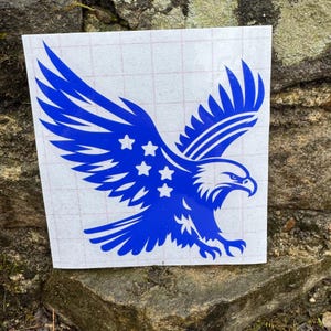 Könnte beinhalten: Ein blauer Adler-Aufkleber mit weißen Sternen auf den Flügeln. Der Aufkleber befindet sich auf einem weißen Blatt Papier mit einem Gittermuster.