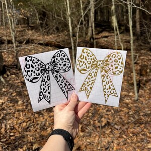 Peut inclure: Deux autocollants carr&eacute;s pr&eacute;sentant un grand motif de n&oelig;ud avec un imprim&eacute; l&eacute;opard. Un autocollant est noir et l'autre est dor&eacute;. Les deux autocollants sont appliqu&eacute;s sur un papier transfert blanc quadrill&eacute; pour faciliter la pose.