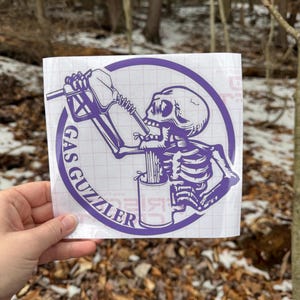 May include: A purple vinyl decal on a white grid backing featuring a skeleton drinking from a gas pump nozzle into a mug. The circular design includes the text GAS GUZZLER printed in a bold, sans-serif font along the left side of the border.