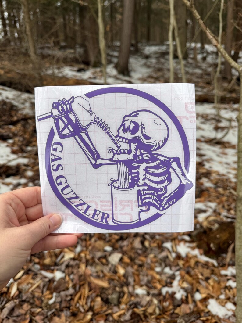 May include: A purple vinyl decal on a white grid backing featuring a skeleton drinking from a gas pump nozzle into a mug. The circular design includes the text GAS GUZZLER printed in a bold, sans-serif font along the left side of the border.