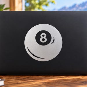 May include: A matte black laptop featuring a large, centered white graphic of a billiard eight ball. The circular design displays the number eight in a bold black font within a white circle, accented by a curved white highlight on the lower edge.