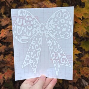Peut inclure: Un autocollant blanc repr&eacute;sentant un grand n&oelig;ud avec un motif l&eacute;opard d&eacute;coup&eacute;. L'autocollant est fix&eacute; sur une feuille de transfert translucide avec un quadrillage rouge clair, con&ccedil;u pour &ecirc;tre appliqu&eacute; sur diverses surfaces lisses.