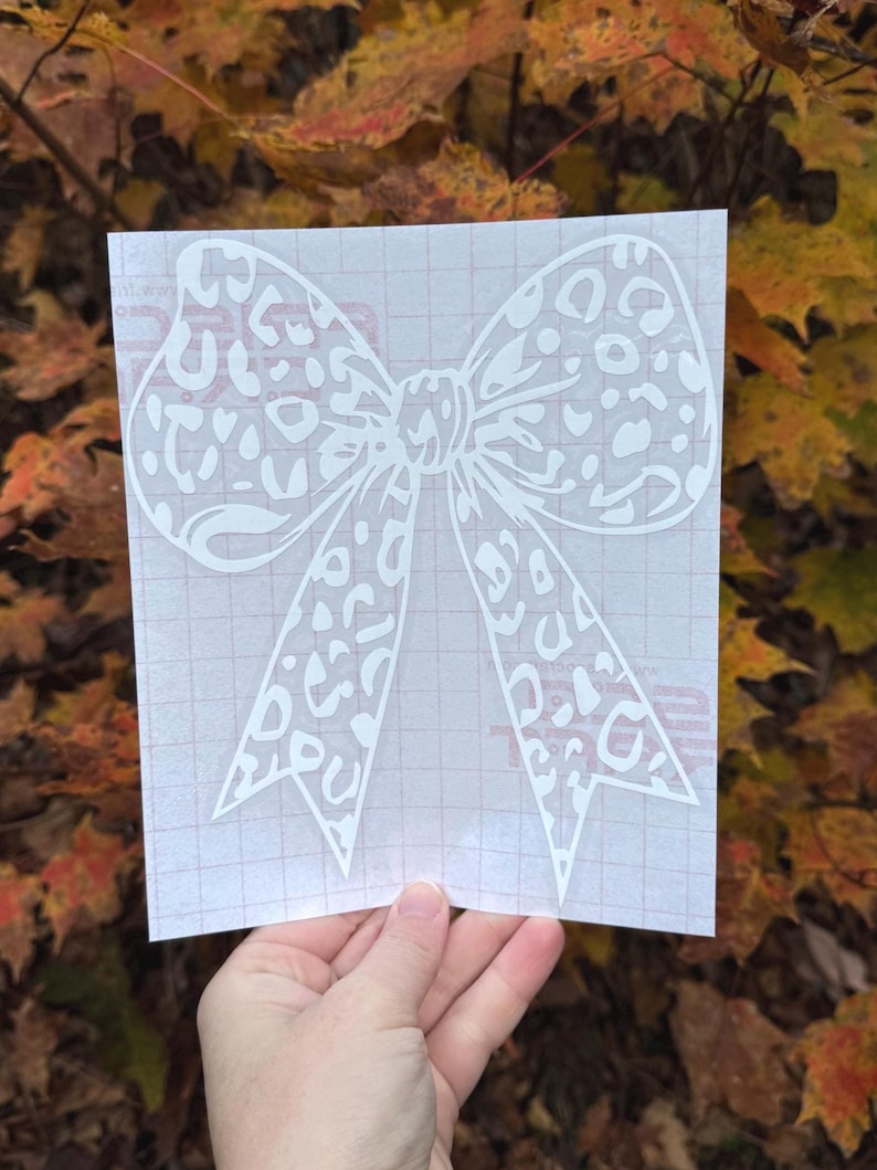Peut inclure: Un autocollant blanc repr&eacute;sentant un grand n&oelig;ud avec un motif l&eacute;opard d&eacute;coup&eacute;. L'autocollant est fix&eacute; sur une feuille de transfert translucide avec un quadrillage rouge clair, con&ccedil;u pour &ecirc;tre appliqu&eacute; sur diverses surfaces lisses.
