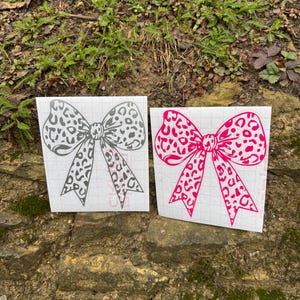 Peut inclure: Deux autocollants en forme de n&oelig;ud avec un motif l&eacute;opard. L'un est gris fonc&eacute; et l'autre est rose vif. Les deux n&oelig;uds sont imprim&eacute;s sur un support en papier transfert blanc quadrill&eacute;, montrant un contour clair du motif.