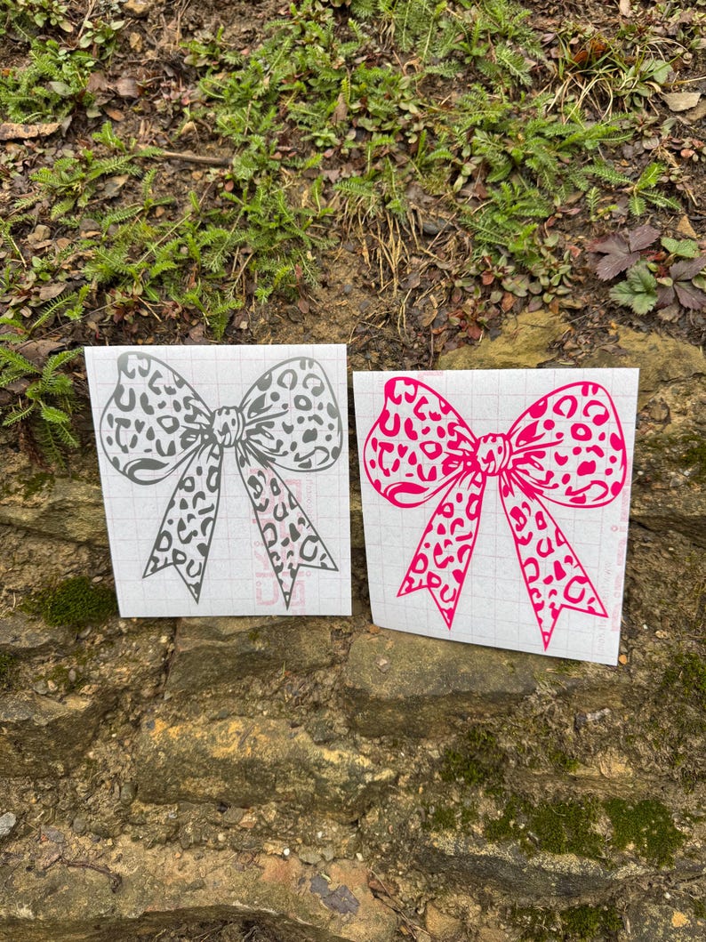 Peut inclure: Deux autocollants en forme de n&oelig;ud avec un motif l&eacute;opard. L'un est gris fonc&eacute; et l'autre est rose vif. Les deux n&oelig;uds sont imprim&eacute;s sur un support en papier transfert blanc quadrill&eacute;, montrant un contour clair du motif.