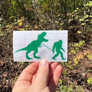 Może przedstawiać: Zielona naklejka winylowa przedstawiająca Tyrannosaurusa Rexa prowadzącego Bigfoota na smyczy. Projekt to humorystyczne spojrzenie na klasyczną dynamikę właściciela i zwierzęcia. Naklejka jest wyświetlana na naturalnym tle zewnętrznym.
