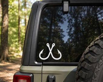 May include: A white vinyl decal featuring two crossed fishing hooks, applied to the rear window of a vehicle. The design is simple and monochromatic, showing the curved shanks and sharp points of the hooks overlapping in a symmetrical arrangement.