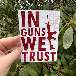 Op de afbeelding: Rode vinyl sticker met de tekst "In Guns We Trust" en een silhouet van een geweer.