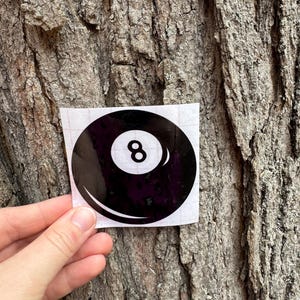 May include: A circular black sticker featuring a white center circle with the number 8 printed in black, designed to resemble a billiard ball. The sticker is presented on a square white backing sheet with a visible grid pattern on the surface.