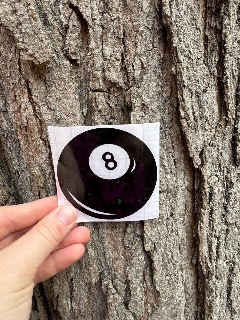 May include: A circular black sticker featuring a white center circle with the number 8 printed in black, designed to resemble a billiard ball. The sticker is presented on a square white backing sheet with a visible grid pattern on the surface.