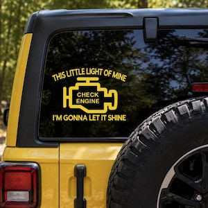 May include: A yellow vinyl decal applied to a vehicle rear window. The design features a stylized check engine icon with the text "THIS LITTLE LIGHT OF MINE" above and "I'M GONNA LET IT SHINE" below, with the words "CHECK ENGINE" inside the icon.