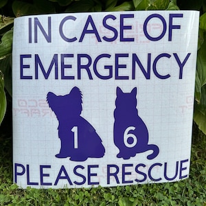 May include: A white sign with dark blue text and graphics. The text reads "IN CASE OF EMERGENCY PLEASE RESCUE." Silhouettes of a dog and cat are featured, with the numbers "1" and "6" inside. The sign is placed on grass.