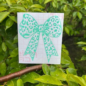 Peut inclure: Un autocollant en vinyle sarcelle en forme de n&oelig;ud avec un motif l&eacute;opard, appliqu&eacute; sur une feuille de support rectangulaire blanche avec un quadrillage rouge p&acirc;le. L'autocollant pr&eacute;sente deux boucles et deux longues queues pointues.