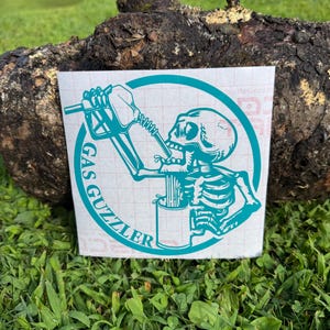 May include: A teal-colored vinyl decal featuring a skeleton drinking from a gas pump nozzle into a mug, enclosed within a circular border. The text GAS GUZZLER is printed in a bold, sans-serif font along the left side of the circular frame.