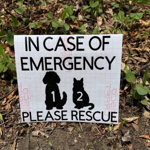 May include: A white sign with black text that reads "IN CASE OF EMERGENCY PLEASE RESCUE." Silhouettes of a dog and cat are below the text, with the numbers 1 and 2 inside. The sign is placed on a bed of leaves and dirt.