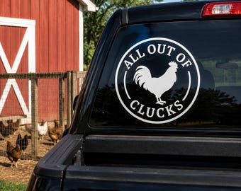 May include: A white circular vinyl decal applied to the rear window of a black truck. The design features a silhouette of a rooster in the center, surrounded by the text ALL OUT OF CLUCKS arranged in a circular border around the central graphic.