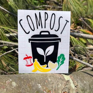 May include: A white sticker with the word "COMPOST" in black script above a black compost bin graphic. The bin features a leaf design and is surrounded by colorful illustrations of food scraps, including a red apple, banana, and lettuce.