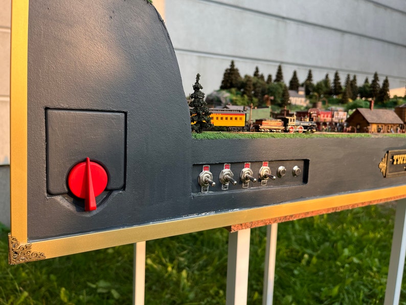 Pu&ograve; includere: Un set di treni in miniatura con una base nera e dorata, una manopola di controllo rossa e cinque interruttori a levetta argentati. Il treno viaggia attraverso una scena di foresta in miniatura.