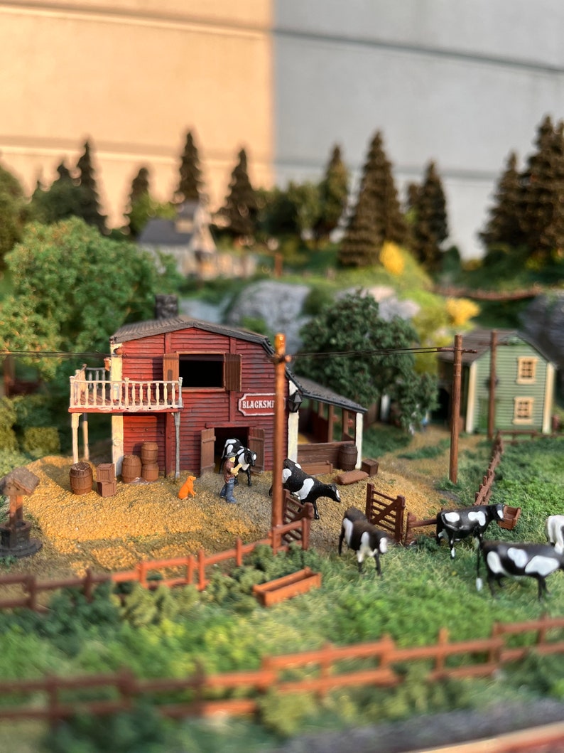 Pu&ograve; includere: Un modello in miniatura di una scena di fattoria con un fienile rosso con l'etichetta "Blacksmith", un piccolo cane e diverse mucche bianche e nere. La scena &egrave; ambientata in un campo erboso con una recinzione di legno e una piccola mangiatoia di legno.