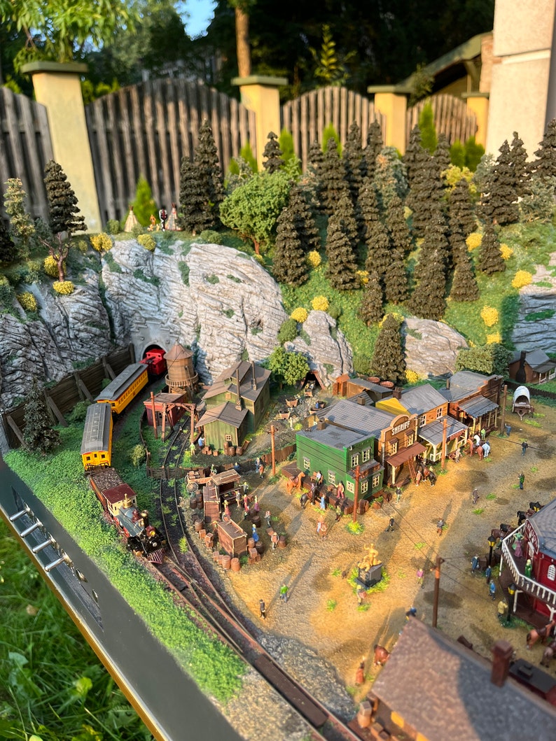 Pu&ograve; includere: Un set di treni in miniatura con una locomotiva a vapore che tira un vagone passeggeri giallo e rosso attraverso un tunnel di montagna. Il set di treni in miniatura &egrave; su un binario che attraversa una citt&agrave; in miniatura con edifici, alberi e figure.